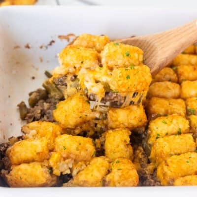 Wooden spoon with tater tot casserole dish with ground beef, green beans, cheddar cheese and golden tater tots.