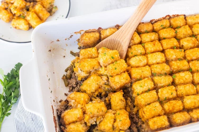 Wooden spoon with tater tot casserole dish with ground beef, green beans, cheddar cheese and golden tater tots.