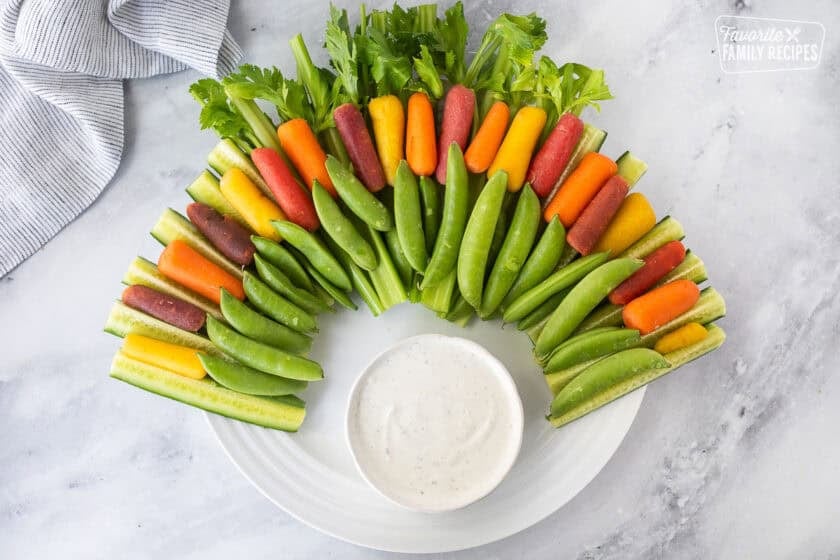 Large round plate with arranged celery stalks, sliced cucumbers, sugar snap peas and colored baby carrots. Small bowl of ranch dip on the bottom of the plate.
