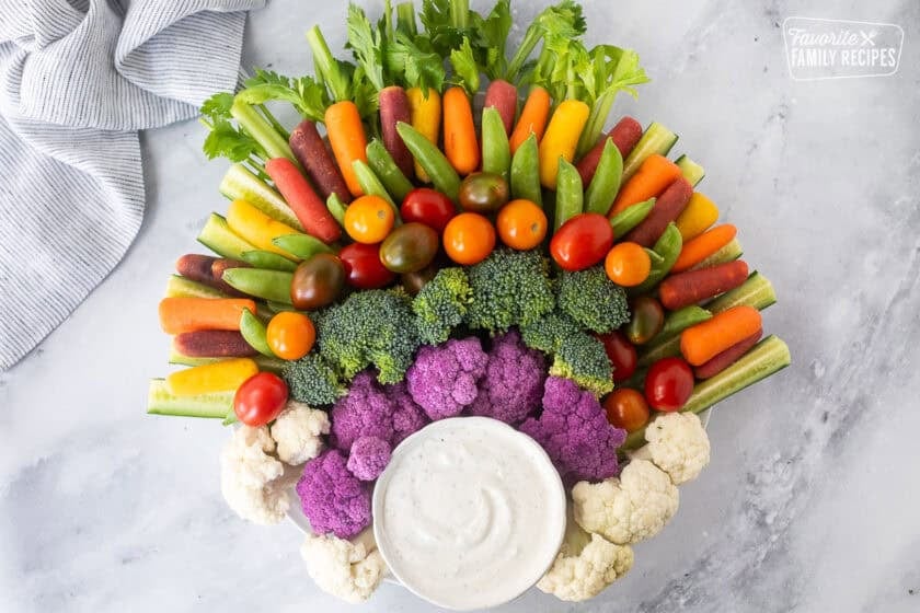 Large round plate with arranged celery stalks, sliced cucumbers, sugar snap peas, colored baby carrots, broccoli, cauliflower and purple cauliflower. Small bowl of ranch dip on the bottom of the plate.