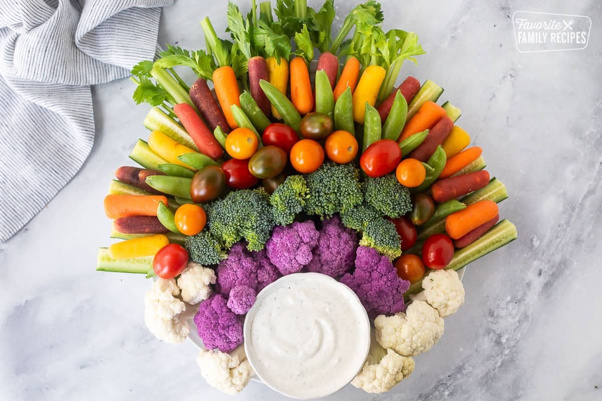 Large round plate with arranged celery stalks, sliced cucumbers, sugar snap peas, colored baby carrots, broccoli, cauliflower and purple cauliflower. Small bowl of ranch dip on the bottom of the plate.