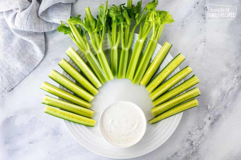 Large round plate with celery stalks and sliced cucumbers arranged at top of the plate. Small bowl of ranch dip on the bottom of the plate.