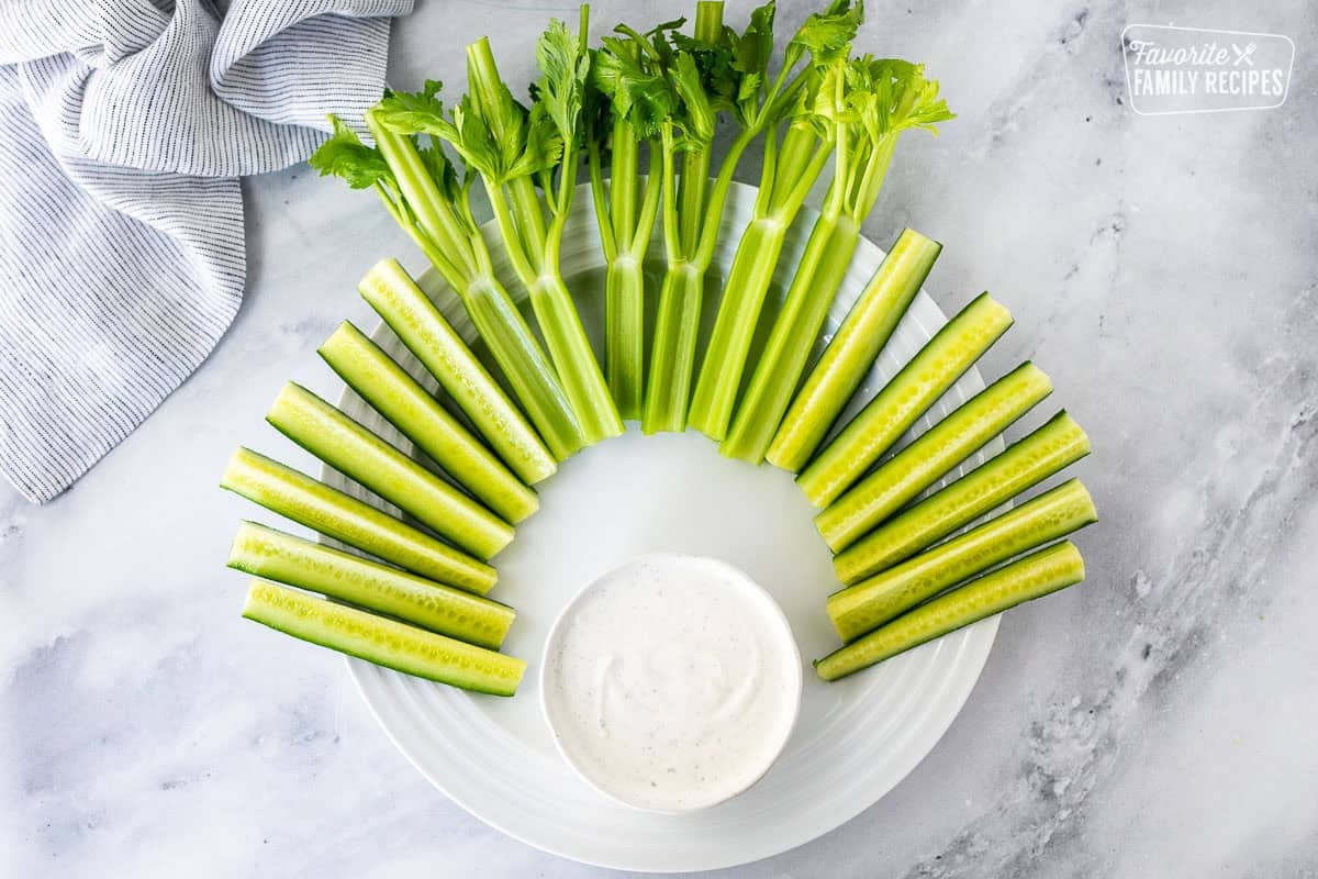 Large round plate with celery stalks and sliced cucumbers arranged at top of the plate. Small bowl of ranch dip on the bottom of the plate.