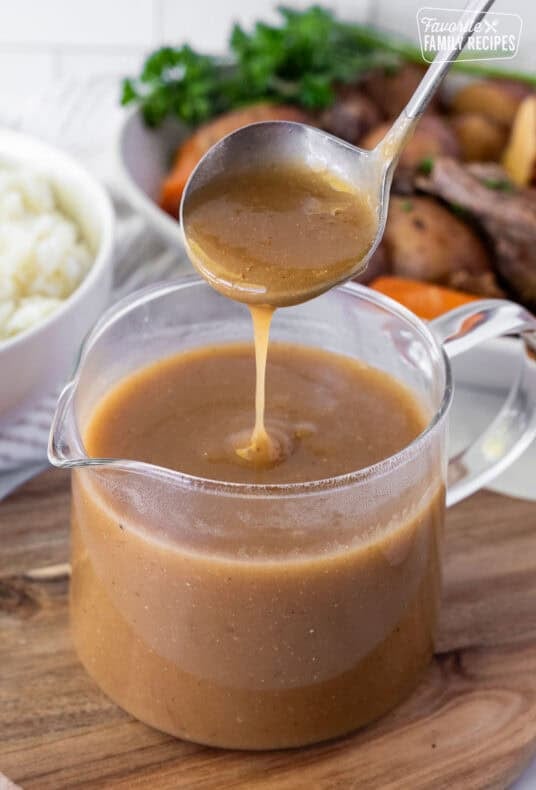 Brown gravy dripping from a ladle into a glass pitcher, with pot roast, carrots, and mashed potatoes in the background.