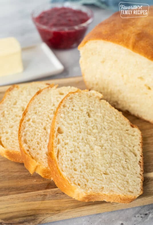 Sliced white bread with a golden crust on a wooden cutting board, with butter and red jam in the background.