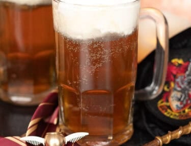 Harry Potter butterbeer topped with creamy foam in glass mugs beside a Gryffindor tie, golden snitch, and wand.