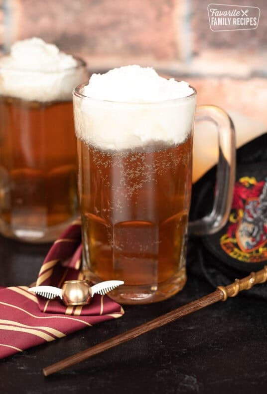 Harry Potter butterbeer topped with creamy foam in glass mugs beside a Gryffindor tie, golden snitch, and wand.