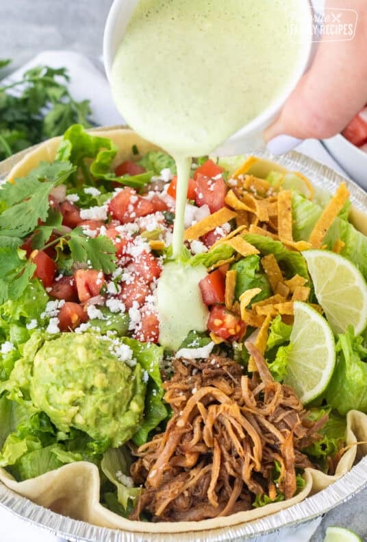 Creamy green dressing poured over a pulled pork taco salad with lettuce, tomatoes, guacamole, cheese, and tortilla strips.