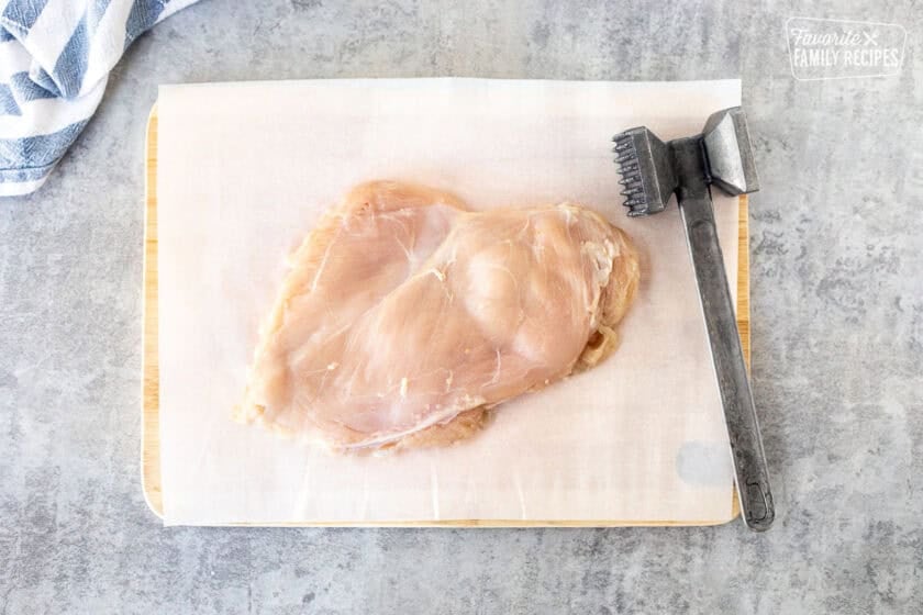 Cutting board covered with parchment paper and thin chicken breast with a mallet on the side.