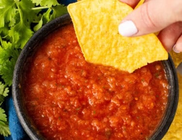 Hand dipping a tortilla chip into tomato salsa in a black bowl, surrounded by cilantro, lime, and jalapeños.