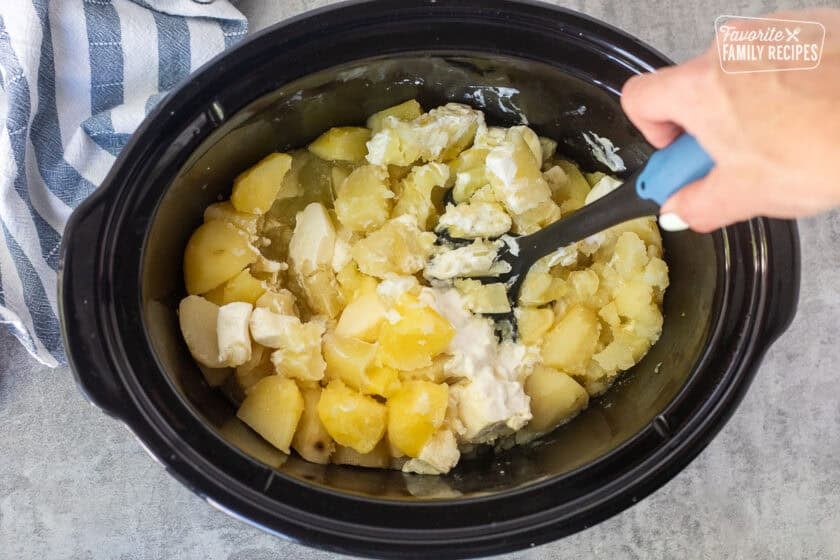Mashing cooked potatoes with butter, sour cream, and cream cheese in the slow cooker.