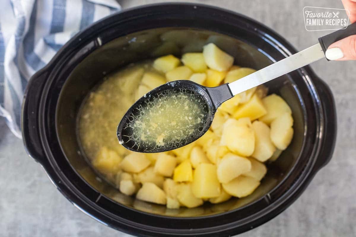 Spooning excess cooking liquid out of the slow cooker before mashing the potatoes.