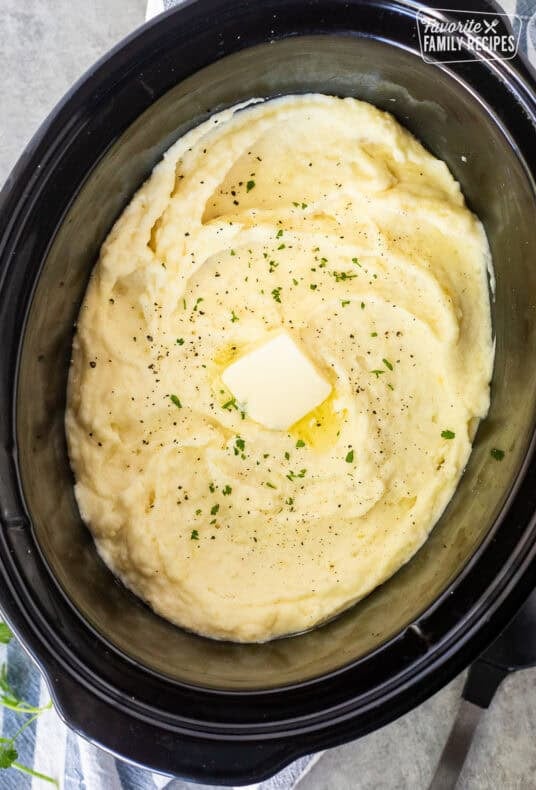 A crockpot filled with mashed potatoes topoed with a square of butter, pepper and fresh parsley.