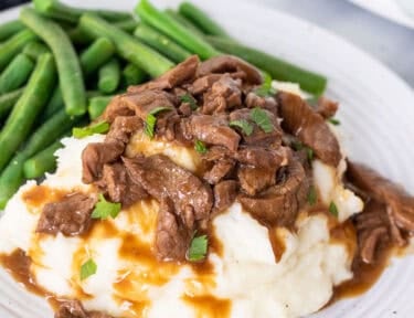 Crockpot steak and gravy served over mashed potatoes, garnished with parsley, with green beans on a white plate.