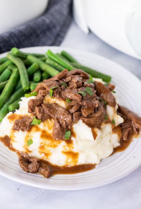 Crockpot steak and gravy served over mashed potatoes, garnished with parsley, with green beans on a white plate.