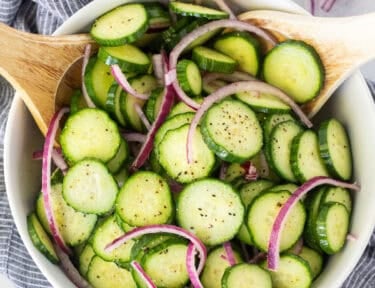 Cucumber salad with thinly sliced red onions and cracked black pepper in a white bowl with wooden serving spoons.