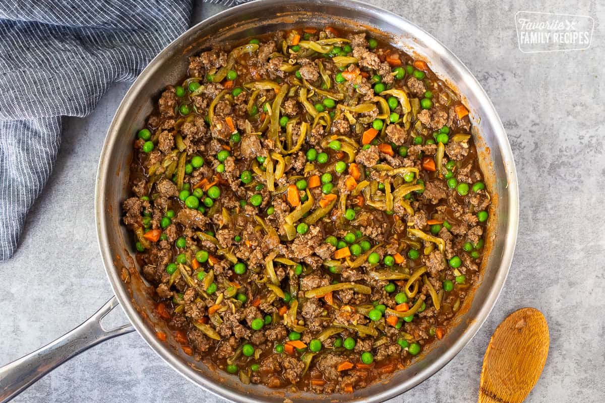 Shepherds pie filling in a skillet including ground beef, tomato sauce, French style green beans, peas, carrots and onion.