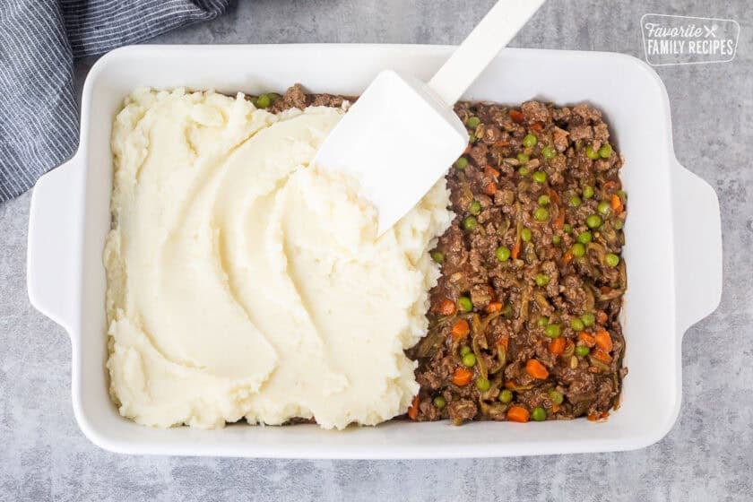 Using a spatula to spread mashed potatoes on top of shepherds pie ground beef mixture.