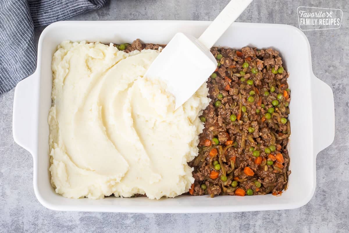 Using a spatula to spread mashed potatoes on top of shepherds pie ground beef mixture.