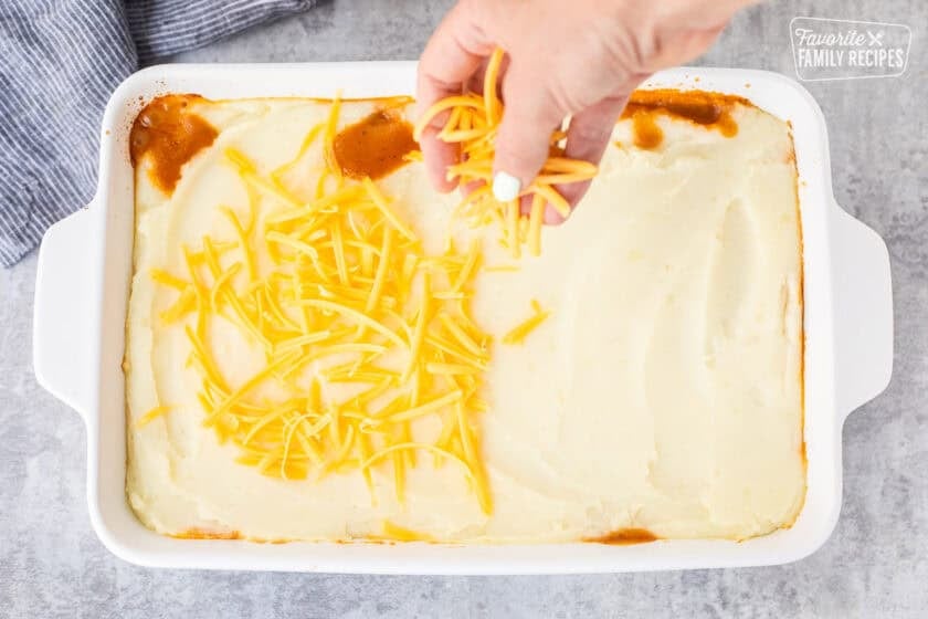 Sprinkling shredded cheddar cheese on top of baked shepherds pie in a casserole dish.