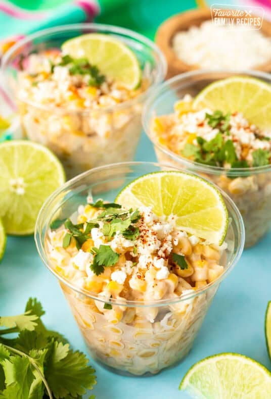 Mexican street corn cups topped with crumbled cheese, cilantro, chili powder, and lime wedges.
