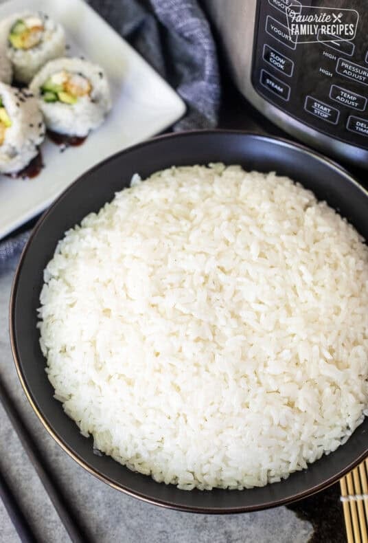 Sushi rice in a black bowl beside an electric pressure cooker, with sushi rolls and a bamboo rolling mat nearby.