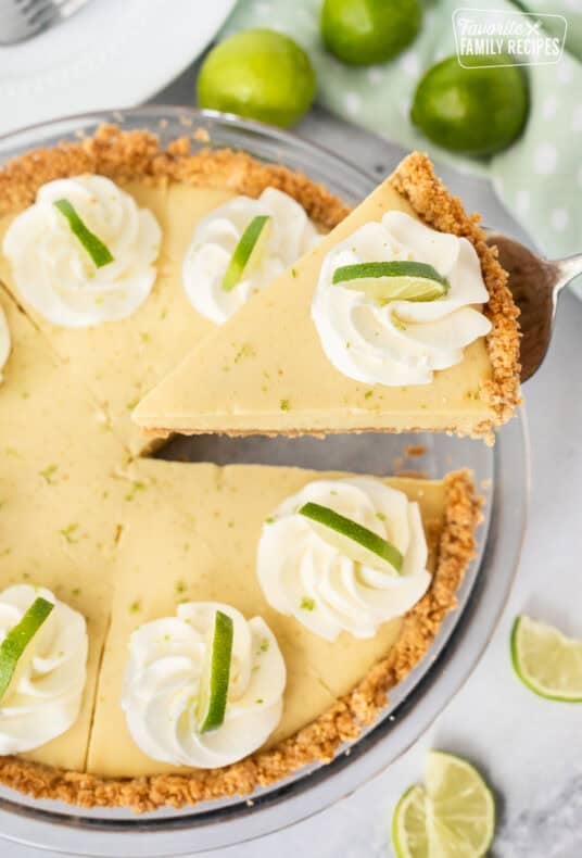 Key lime pie with a graham cracker crust, whipped cream, and lime slices, with one slice lifted on a pie server.