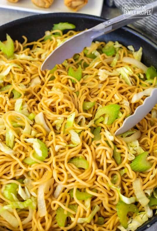 Vegetable chow mein noodles with cabbage, celery, and onions in a black skillet with serving tongs.