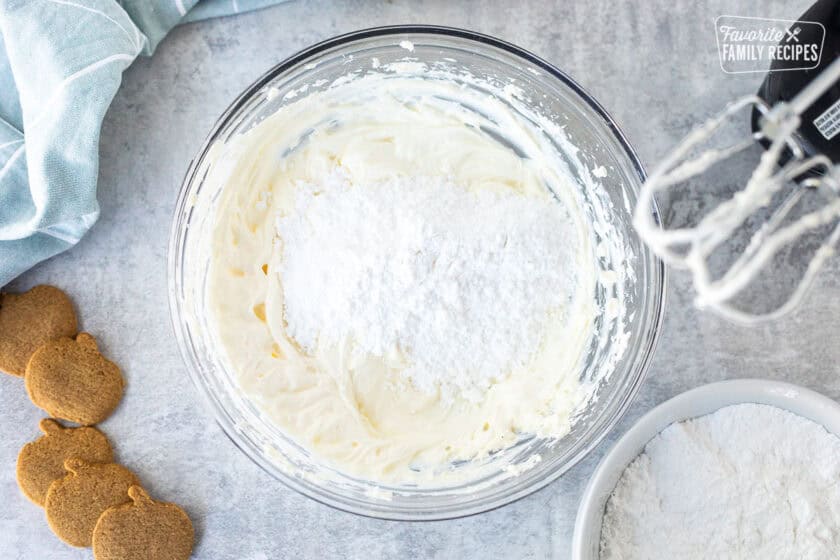 Glass mixing bowl with whipped cream cheese, sour cream and little bit of powdered sugar. Electric mixer on the side and bowl of powdered sugar.