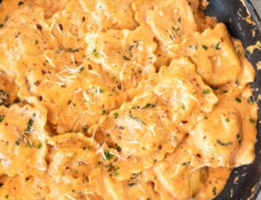 Ravioli in creamy tomato sauce topped with shredded Parmesan, herbs, and red pepper flakes in a black skillet.