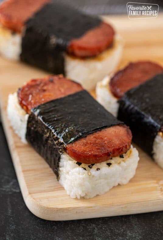 Spam musubi with browned Spam slices over white rice, wrapped in nori seaweed and served on a wooden board.