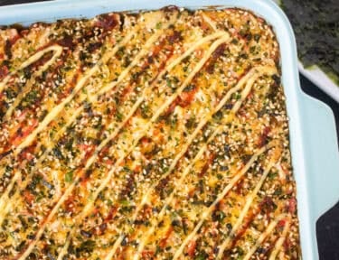 Sushi bake topped with creamy sauce, sesame seeds, and seaweed flakes in a blue baking dish, with nori and soy sauce alongside.
