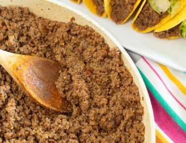 Taco Bell copycat seasoned ground beef in a skillet beside crunchy tacos topped with shredded lettuce and cheddar cheese.
