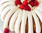 White chocolate raspberry Bundt cake with thick piped frosting, fresh raspberries, and white chocolate pieces on a white platter.