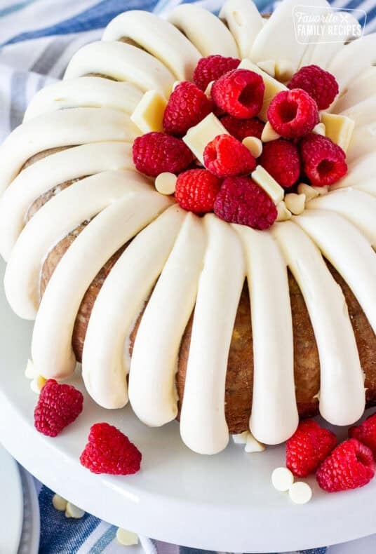 White chocolate raspberry Bundt cake with thick piped frosting, fresh raspberries, and white chocolate pieces on a white platter.